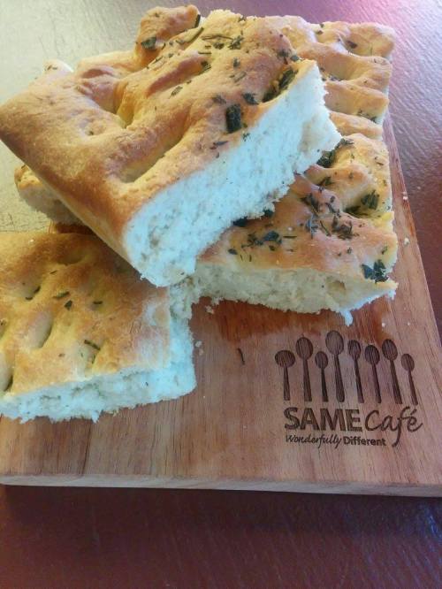 Freshly baked focaccia bread topped with herbs, served on a wooden board with the SAME Café logo.