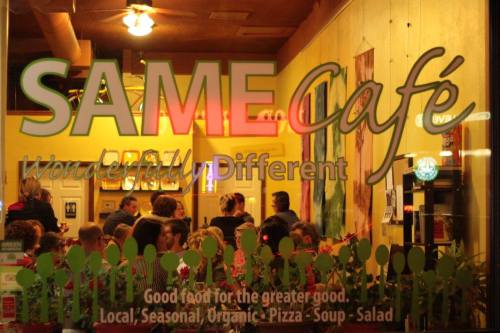 A vibrant café window with the text "SAME Café" and a crowd enjoying food inside, promoting local, organic dishes.