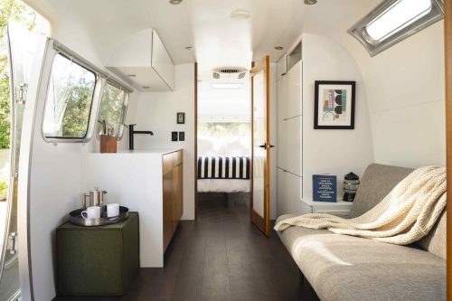 Interior of a modern, minimalist trailer featuring a kitchenette, seating area, and a cozy bedroom in the background.