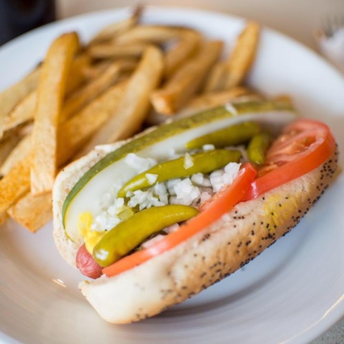 A hot dog in a poppy seed bun topped with pickles, onions, and tomato, served with a side of golden French fries.