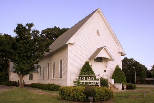 A white Presbyterian church with a peaked roof, surrounded by trees and a sign reading "Oakland Presbyterian."