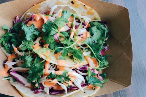 A taco filled with shredded cabbage, purple cabbage, cilantro, and drizzled with a creamy sauce in a brown paper container.