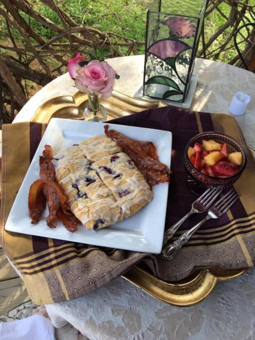 A beautifully arranged breakfast plate with pastries, bacon, and a fruit bowl, set on a table with a rose and decorative lantern.