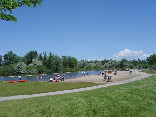 A sunny park scene with people swimming and relaxing by a river, surrounded by green trees and a clear blue sky.