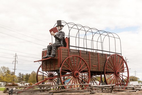 A large statue of a man in a top hat sitting on a wooden wagon, set against a cloudy sky.