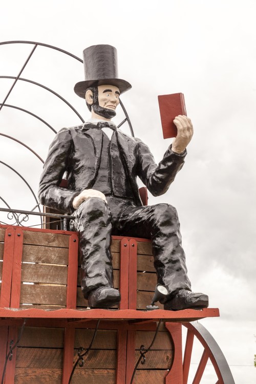 A large statue of a man in a top hat, sitting on a wagon and holding a book, against a cloudy sky.
