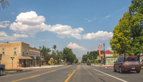 A quiet street scene with shops, trees, and a blue sky dotted with clouds.