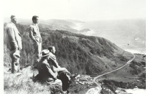 A group of men stands on a cliff overlooking a coastal landscape with a winding road and ocean in the background.