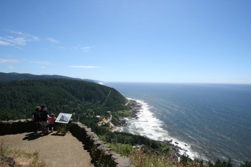 A scenic coastal view with two people observing from a viewpoint, surrounded by lush green hills and ocean waves.