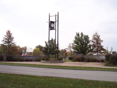 A clock tower surrounded by trees and landscaped gardens, with a road in the foreground and flags in the background.
