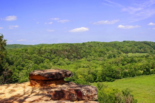 A rocky outcrop overlooks a lush green valley under a blue sky with scattered clouds.