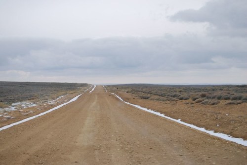 A dirt road stretches into the distance, flanked by sparse vegetation under a cloudy sky.