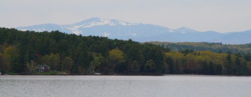 A serene lake surrounded by lush trees, with snow-capped mountains in the background under a cloudy sky.