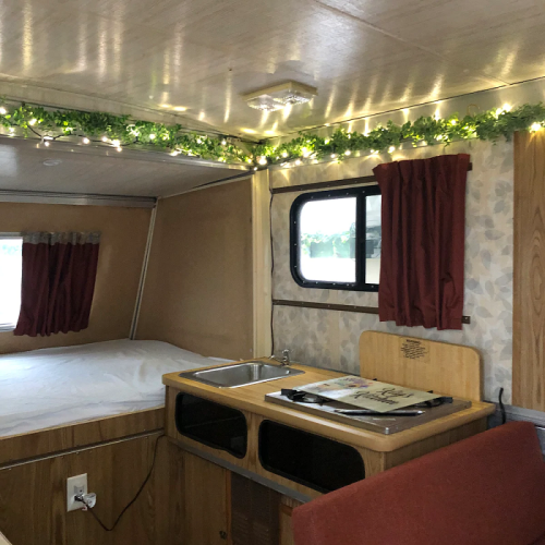 Cozy interior of a camper with a bed, kitchenette, and decorative lights along the ceiling.