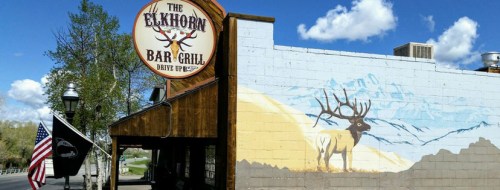 Exterior of The Elkhorn Bar & Grill featuring a mural of a deer and mountains, with an American flag nearby.