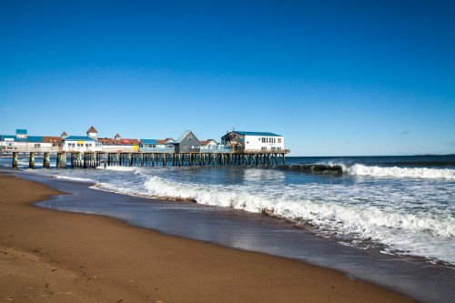A wooden pier extends over a sandy beach, with waves gently crashing and a clear blue sky above.