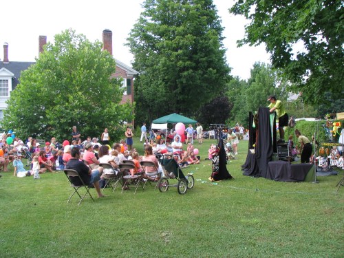 A crowd of children and adults watching a performance outdoors, surrounded by trees and colorful decorations.