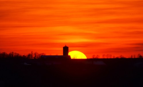 Silhouette of a barn and silo against a vibrant orange sunset sky.