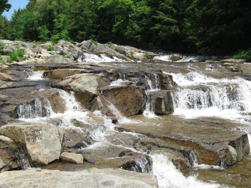 A serene river cascades over rocky terrain, surrounded by lush green trees under a clear blue sky.