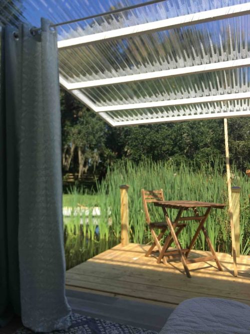 A wooden deck with a table and chair, surrounded by tall green grass and a clear blue sky above.