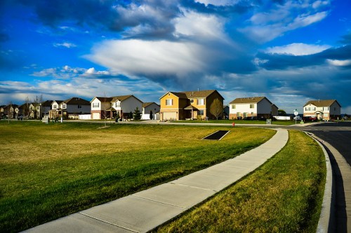 A suburban neighborhood with houses lining a grassy area under a blue sky with clouds, featuring a sidewalk in the foreground.