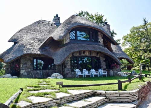 A charming stone cottage with a thatched roof, surrounded by greenery and featuring a stone pathway and outdoor seating.