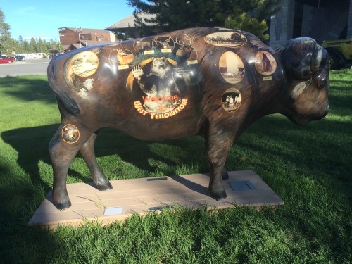 A painted bison statue with various images and text, set on grass near a building in Yellowstone.