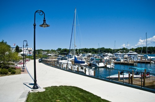 A sunny marina with sailboats and yachts docked, surrounded by a walkway and green grass under a clear blue sky.