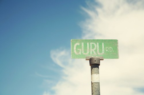 Green street sign reading "GURU RD." against a blue sky with wispy clouds.