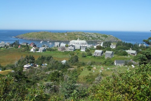 A scenic view of a coastal village with houses, greenery, and boats near an island under a clear blue sky.