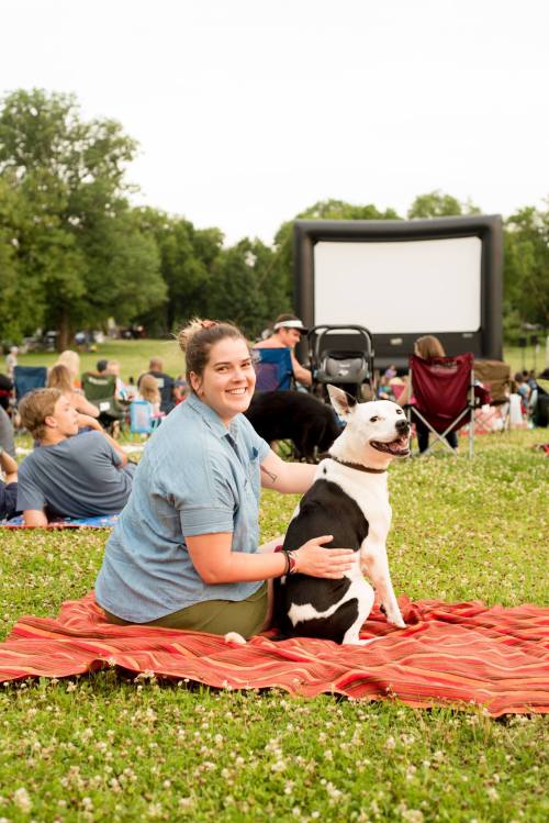 A woman sits on a blanket with her dog, smiling at the camera, while people enjoy a movie in the background.