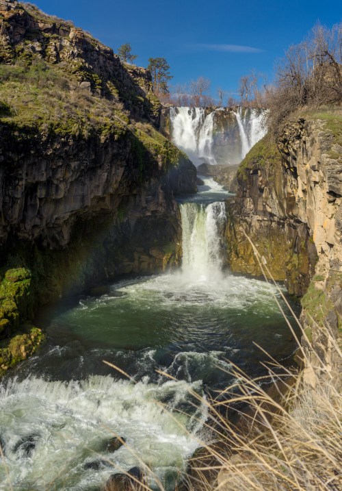 A scenic view of cascading waterfalls surrounded by rocky cliffs and lush greenery under a clear blue sky.