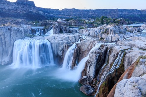A scenic view of a waterfall cascading over rocky cliffs into a turquoise river, surrounded by hills and vegetation.