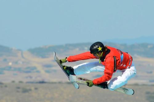 A snowboarder in a red jacket performs a mid-air trick against a clear blue sky and distant landscape.