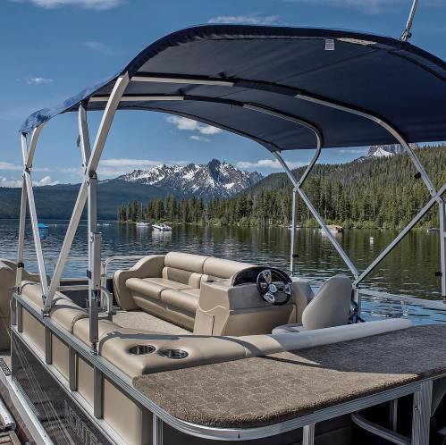A spacious pontoon boat with a canopy, docked on a serene lake surrounded by mountains and lush trees.