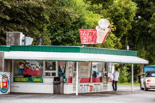 A colorful ice cream stand named "Tasty Dip" with a neon sign, surrounded by trees and a customer at the counter.