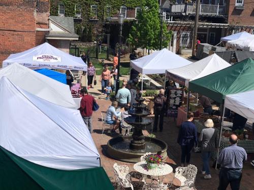 A bustling outdoor market scene with tents, vendors, and visitors around a central fountain on a sunny day.