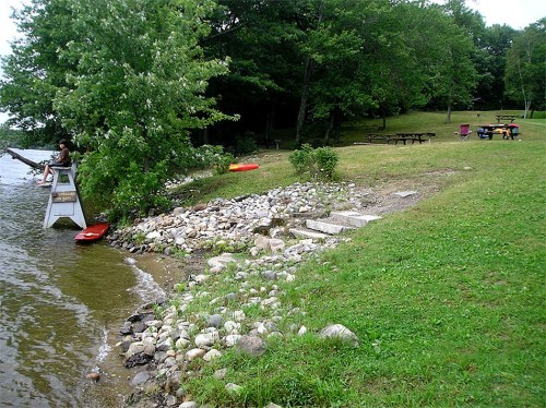 A lakeside scene with a person on a dock, picnic tables, and kayaks near a grassy area and trees.