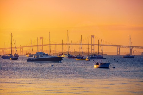 A serene sunset over a harbor filled with sailboats, with a bridge silhouetted in the background.