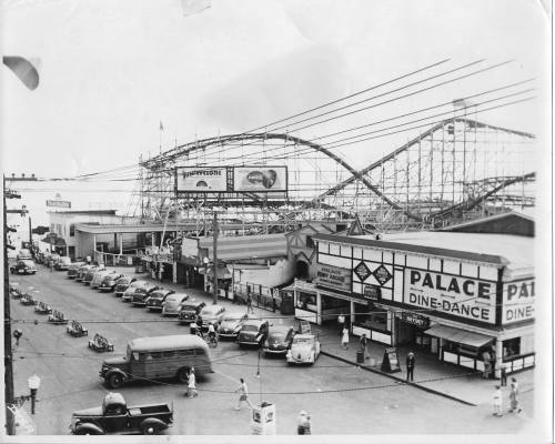Historic black-and-white photo of a busy amusement park street with vintage cars and a roller coaster in the background.