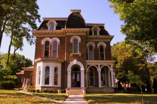A large, historic brick house with ornate windows and a steep roof, surrounded by trees and a clear blue sky.