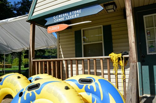 A closed general store with a canoe livery sign, featuring yellow inflatable tubes and a rope on the porch.
