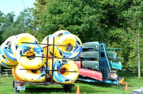 Colorful inflatable tubes stacked on a trailer, with canoes arranged on a rack nearby, set in a green park.
