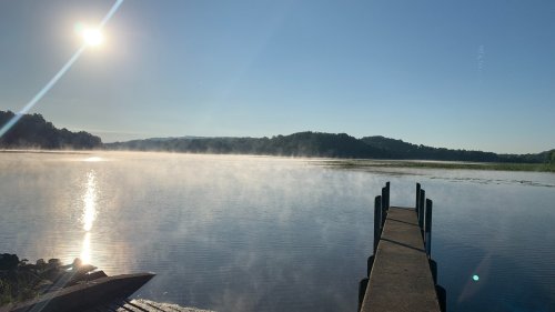 A serene lake at sunrise, with mist rising from the water and a wooden dock extending into the calm scene.
