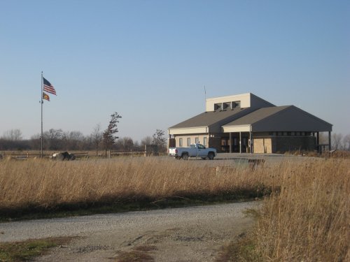 A modern building with a flagpole and American flag, surrounded by tall grass and a gravel road.