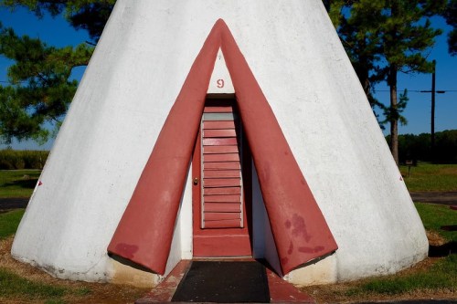 A triangular-shaped building with a red door, set against a clear blue sky and green trees.