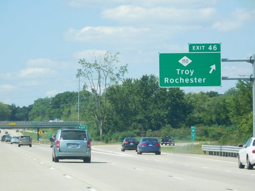 Highway sign indicating exit 46 for Troy and Rochester, with cars traveling on the road under a clear blue sky.