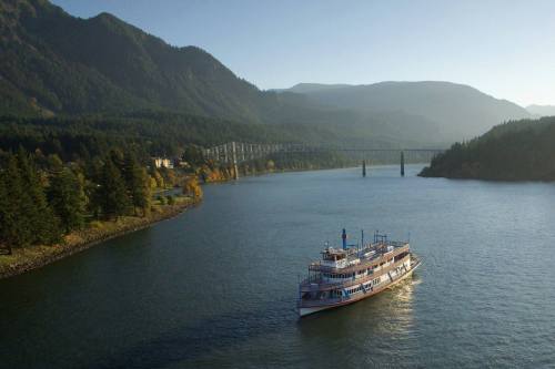 A scenic river view with a boat cruising, surrounded by mountains and a bridge in the background.