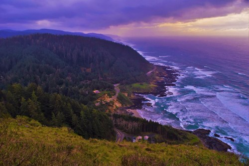 A scenic coastal view with rolling hills, lush forests, and waves crashing against rocky shores under a colorful sky.
