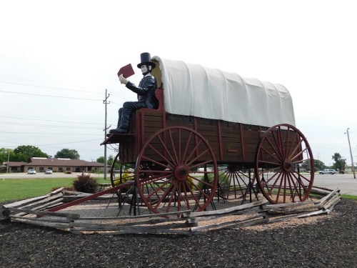 A large statue of a man in a top hat holding a book, sitting on a covered wagon, surrounded by wooden fencing.